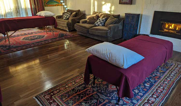 The interior of the Marysville Acupuncture Services treatment room. Two massage tables draped in comfortable cotton blankets with lush pillows sit on decorative rugs. There is plush seating, and everything is lit by dappled sunlight. A cozy fire is burning in the background. Everything is ready for you to relax into healing when you begin using our services!